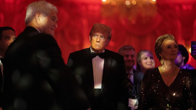 A man in a black tuxedo and bow tie stands amid a formally dressed crowd in a red-lit ballroom, with a woman in a sequined gown beside him.
