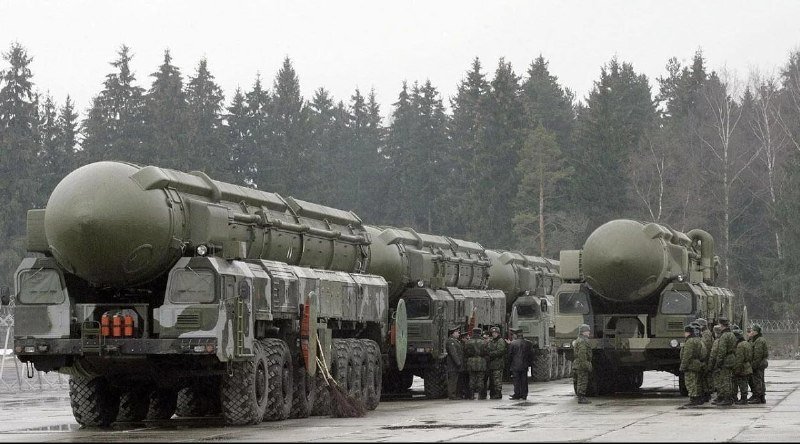 Military personnel in camouflage uniforms stand beside a convoy of large green missile launcher vehicles parked on a paved surface, with a dense evergreen forest in the background.