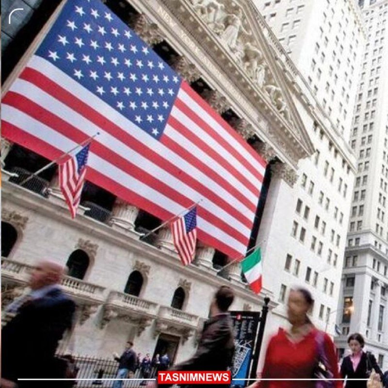 A large American flag and a smaller Italian flag hang from a neoclassical building's facade as pedestrians walk past on the sidewalk below.