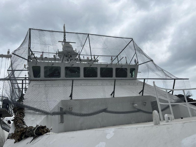 The white upper superstructure and bridge of a naval ship are covered by black netting under an overcast sky.