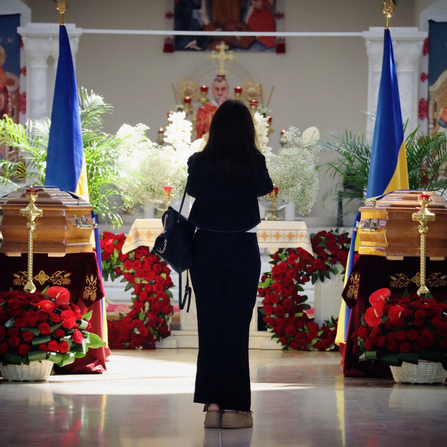 A woman in dark clothing stands facing an altar inside a church, flanked by two wooden coffins draped in flags and surrounded by red floral arrangements.