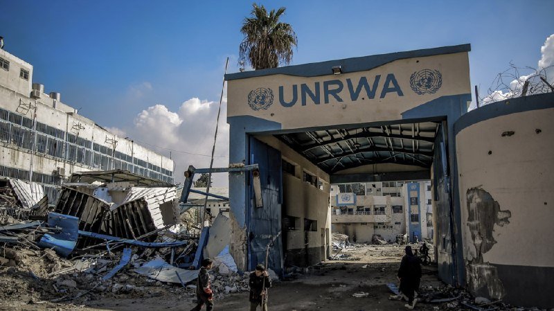 A damaged UNRWA facility stands amid rubble and debris, with two boys and an adult visible near the entrance under a partly cloudy sky.