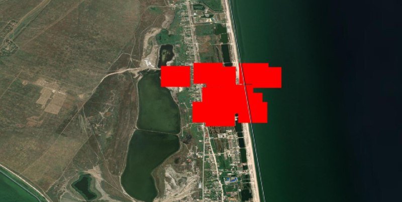 Satellite image showing a coastal area with green fields, inland water bodies, and a developed shoreline, with portions obscured by red rectangular redaction marks.