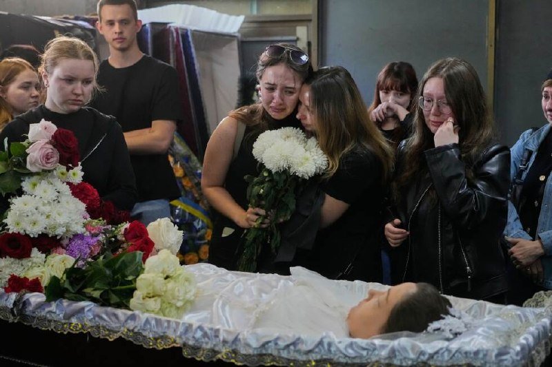 Mourners, some crying and embracing, gather around an open casket holding a person in white, with floral arrangements placed nearby.