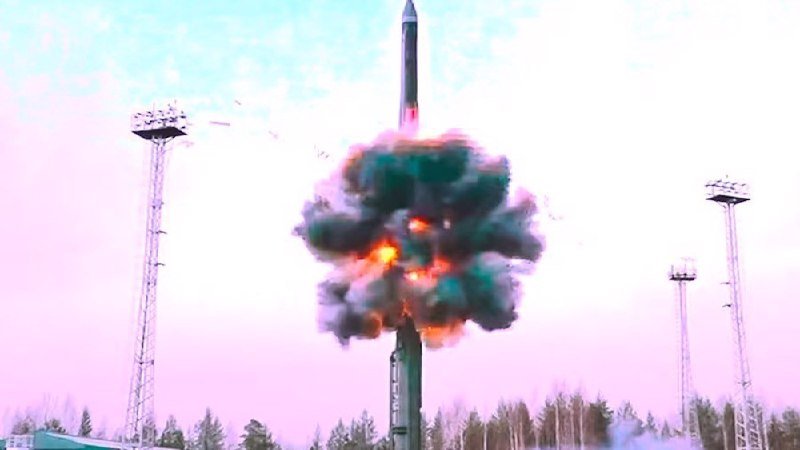 A missile launches upward, engulfed in flames and thick smoke, framed by tall floodlight towers and a treeline against a pale sky.