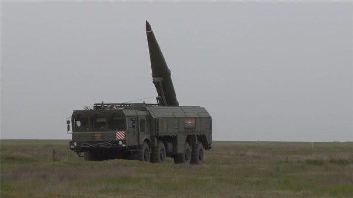 A green military missile launcher vehicle with a raised missile sits on a grassy field under a gray, overcast sky.