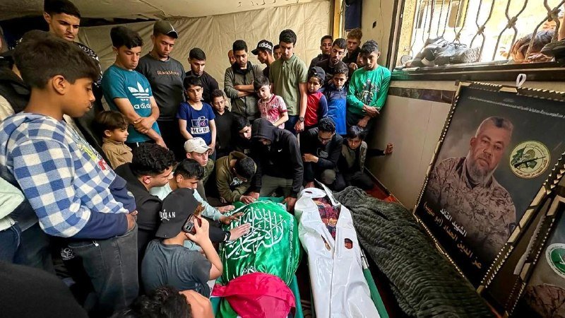 Mourners gather around a green-draped body inside a tent, with a framed portrait of a man displayed nearby.