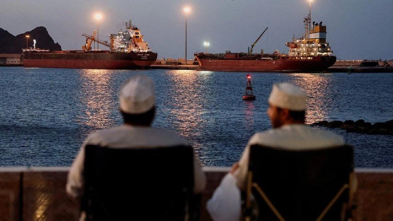 Two men wearing white caps sit in chairs facing a harbor at dusk, where two large cargo ships are anchored and illuminated against a dark hillside.
