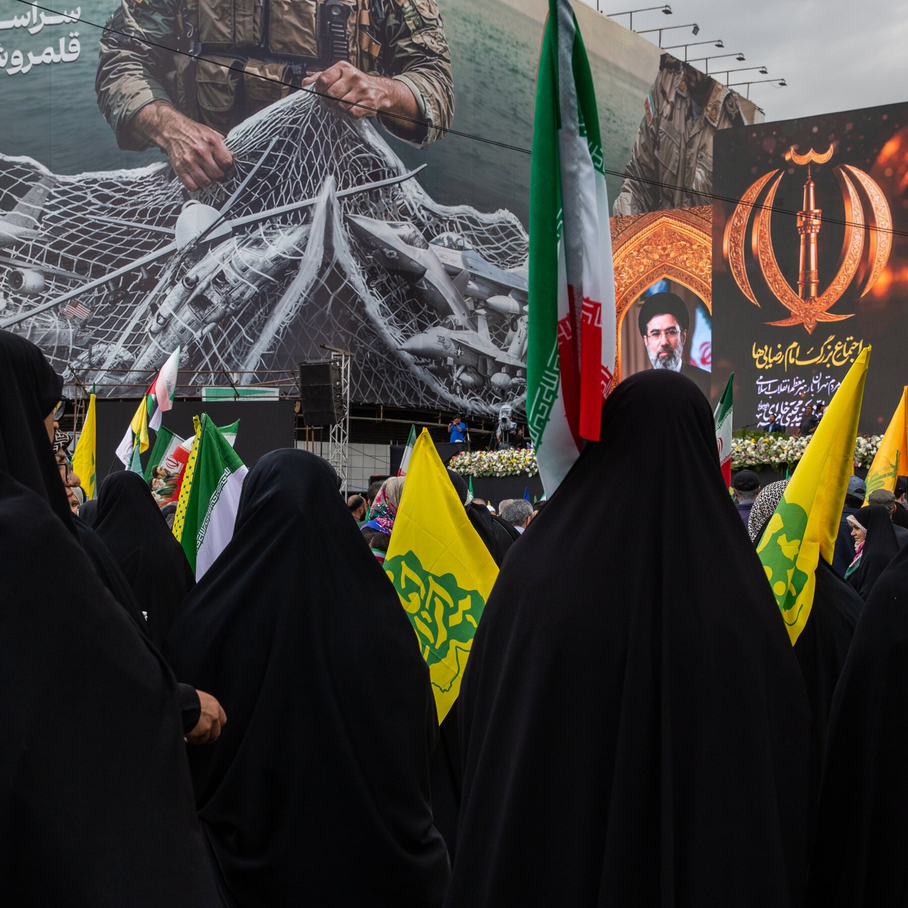 A crowd of women in black chadors holds Iranian and yellow flags, gathered before a stage displaying large military banners with Persian script and a portrait.