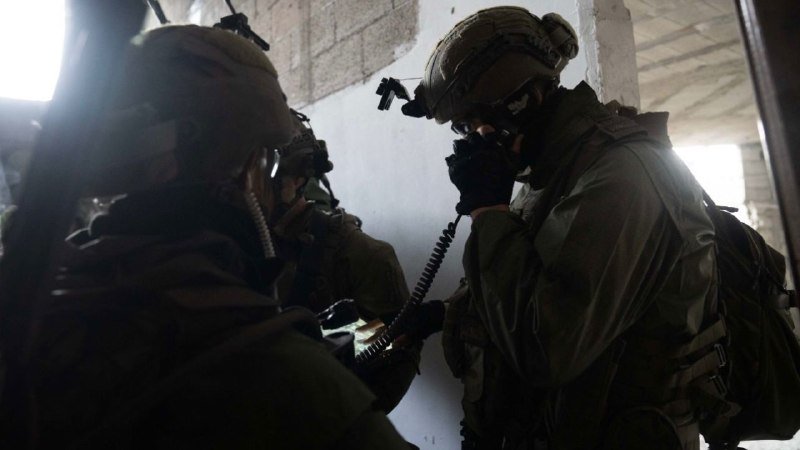Soldiers in tactical gear and helmets speak into coiled radio microphones inside a dimly lit concrete structure.