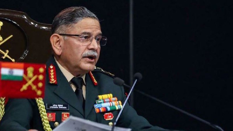 A man in a decorated military uniform with insignia and ribbons speaks at a podium equipped with microphones against a dark background.