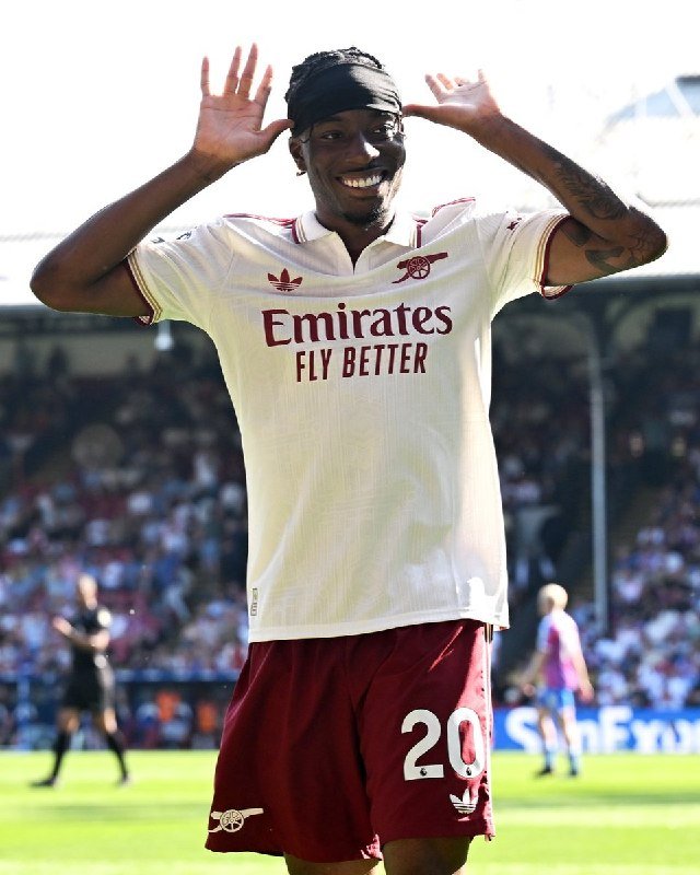 A smiling soccer player wearing a white and burgundy "Emirates Fly Better" jersey with number 20 raises both hands near his head while celebrating on a stadium field.