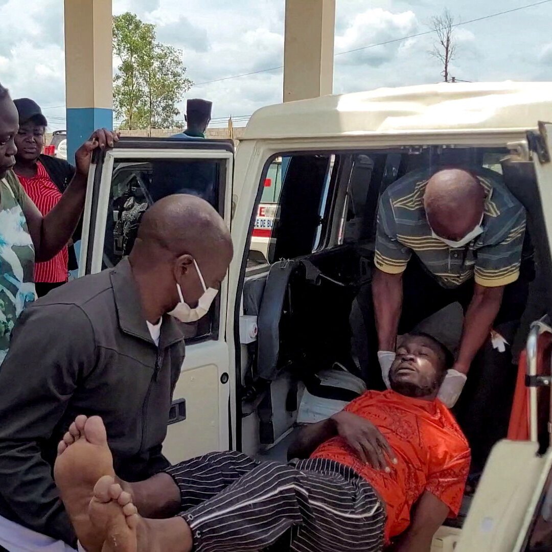 Two masked men lift an injured person in an orange shirt into the back of a white vehicle.