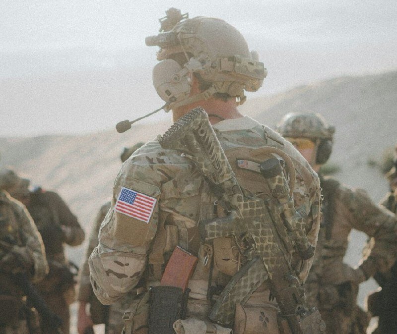 Soldiers in camouflage uniforms and helmets, with one displaying a U.S. flag patch, stand in a mountainous, arid landscape.