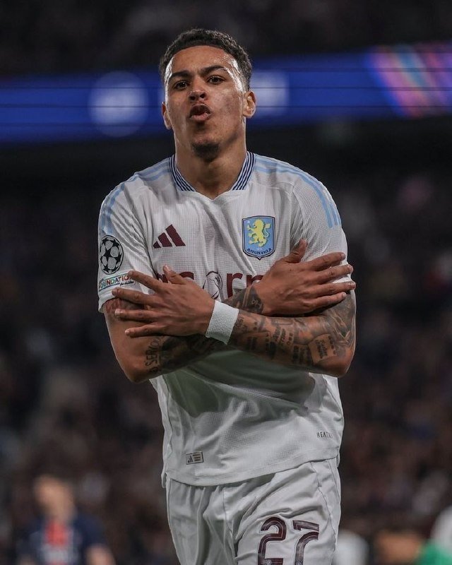 A tattooed soccer player in a white Aston Villa jersey crosses his arms in a goal celebration during a Champions League match.