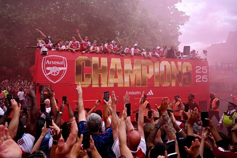 Arsenal football players in red jerseys wave from atop a red double-decker bus displaying "CHAMPIONS 25 26" amid red smoke, as a crowd holds up phones.