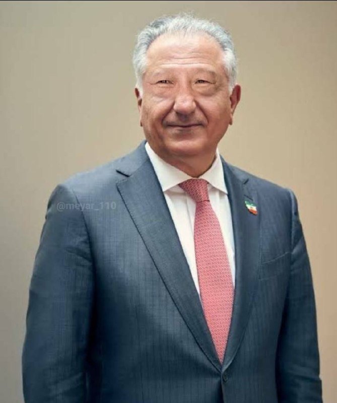 A gray-haired man in a blue suit and pink tie smiles while standing against a plain beige background, wearing a small flag pin on his lapel.
