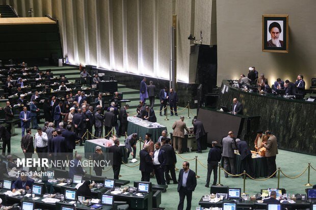 Lawmakers gather on the chamber floor near voting stations during a parliamentary session, with a framed portrait displayed on the wall.