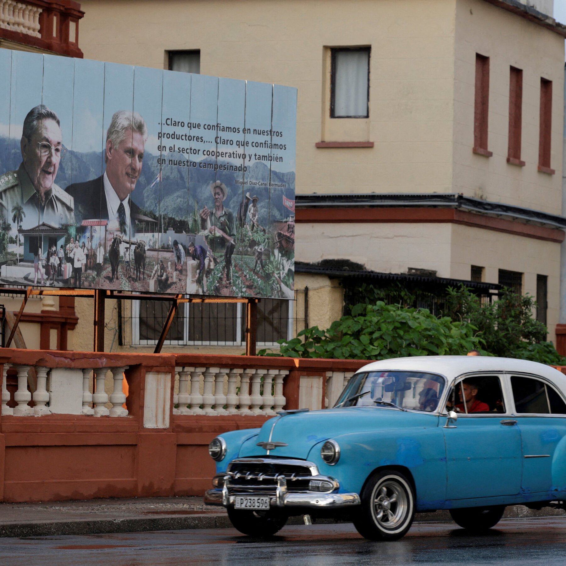 A vintage blue and white car drives past a building displaying a large billboard featuring two men's portraits, Spanish text, and rural imagery.