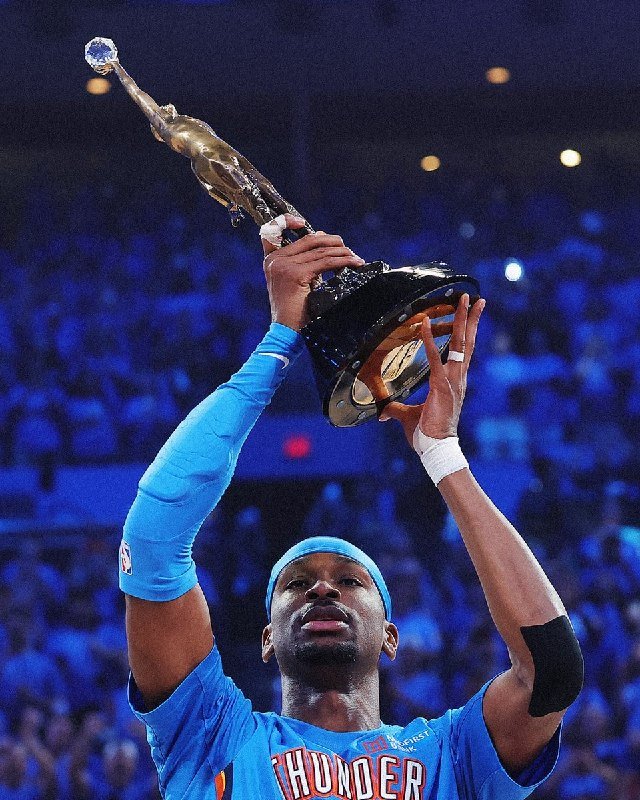 An Oklahoma City Thunder basketball player in a blue jersey and headband hoists a tall silver-and-black trophy above his head in a crowded arena.