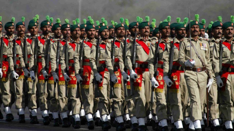 Soldiers in khaki uniforms with red sashes, white gloves, and green berets march in formation while carrying rifles.