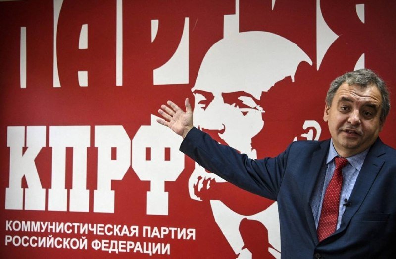 A man in a suit gestures in front of a red banner displaying a stylized portrait and Cyrillic text reading "КПРФ Коммунистическая партия Российской Федерации."