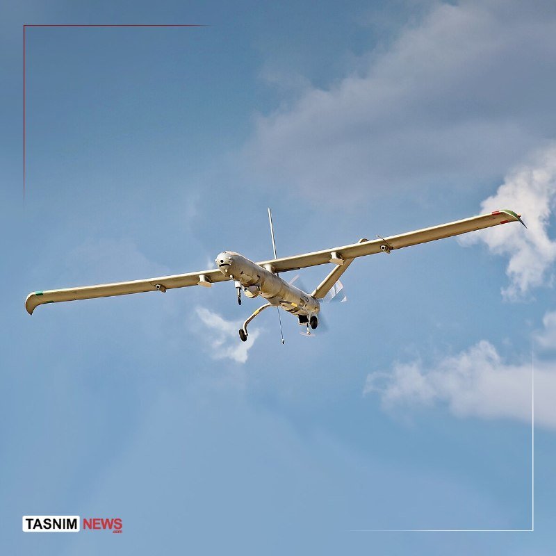 A tan unmanned aerial vehicle with long wings flies against a partly cloudy blue sky, with a "Tasnim News" logo in the corner.