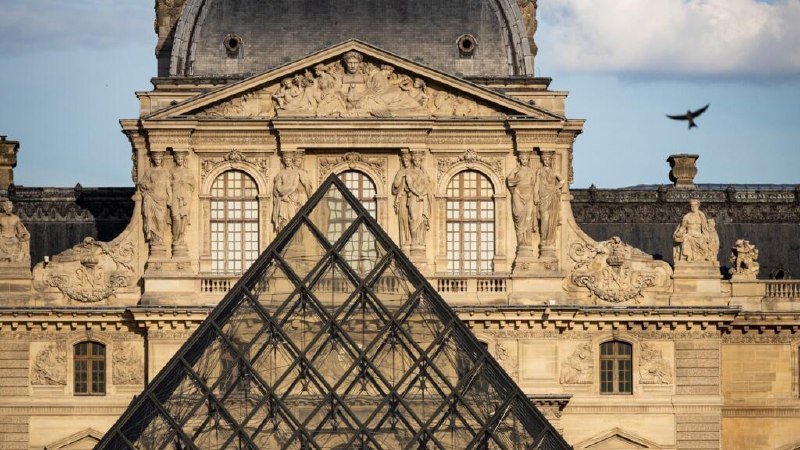 A glass pyramid stands before an ornate stone building featuring classical sculptures, arched windows, and a domed roof under a partly cloudy sky.
