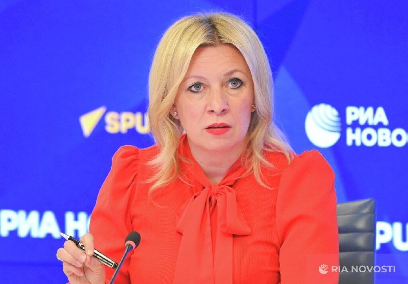 A blonde woman in a red blouse speaks into a microphone against a blue backdrop displaying "РИА НОВОСТИ" and "SPUTNIK" logos.
