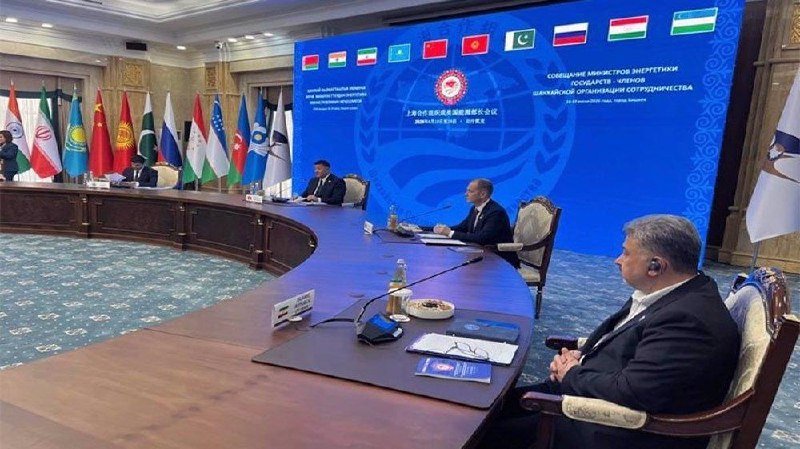 Officials wearing headsets sit at a large curved conference table with microphones and documents, backed by national flags and a blue screen displaying multiple country flags and text.