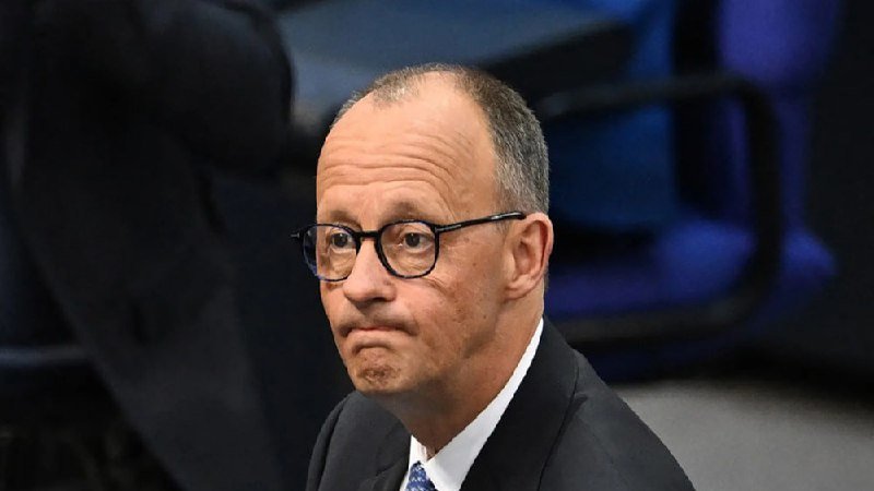 A man in a dark suit, white shirt, and glasses sits with pursed lips in what appears to be an auditorium with blue seating in the background.