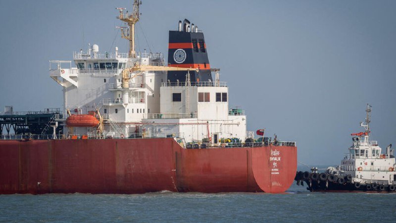 A large red and white cargo ship labeled "DAN.LK" floats on calm water, with a smaller white tugboat positioned to its right.