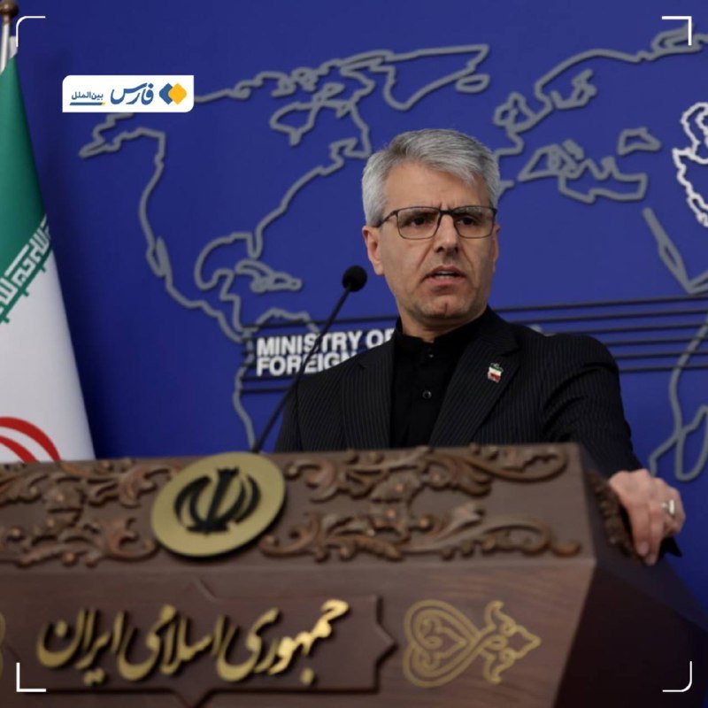 A gray-haired man with glasses speaks at an ornate wooden podium bearing Persian script, with the Iranian flag and a blue "Ministry of Foreign Affairs" world map backdrop behind him.
