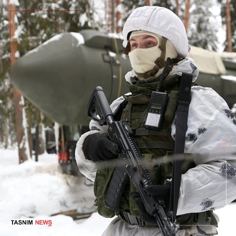 A soldier in white winter camouflage and a helmet stands holding a rifle, with a large green military vehicle visible in the snowy background.