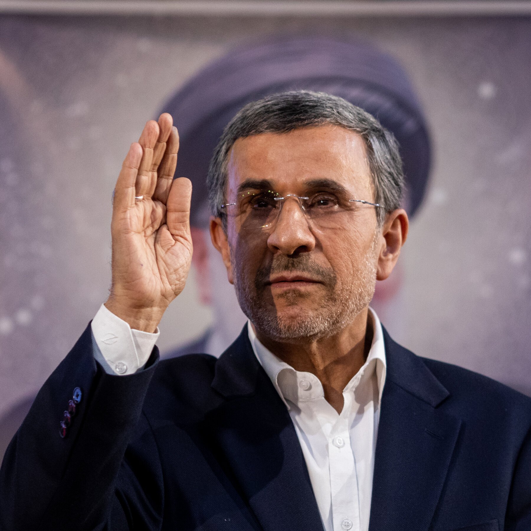 A man with gray hair, glasses, and a beard, wearing a dark blazer and white shirt, raises his right hand in a wave.