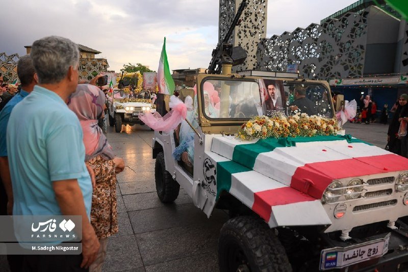 A decorated jeep adorned with flowers, an Iranian flag, and a framed portrait of a cleric drives through a public square as onlookers watch.