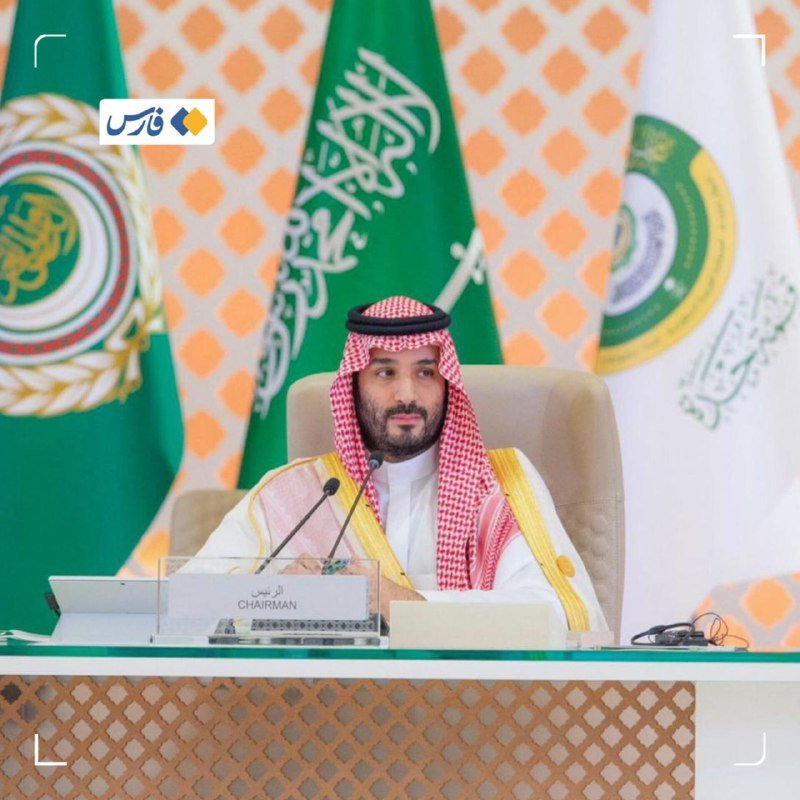 A man wearing a red-checkered keffiyeh and traditional attire sits at a desk with a "CHAIRMAN" nameplate, backed by green and white flags bearing Arabic script.