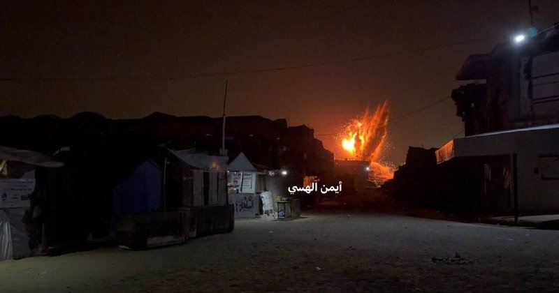 A nighttime street scene shows a large fire burning in the distance beyond small structures, with Arabic text "أمن ألهوى" overlaid on the image.