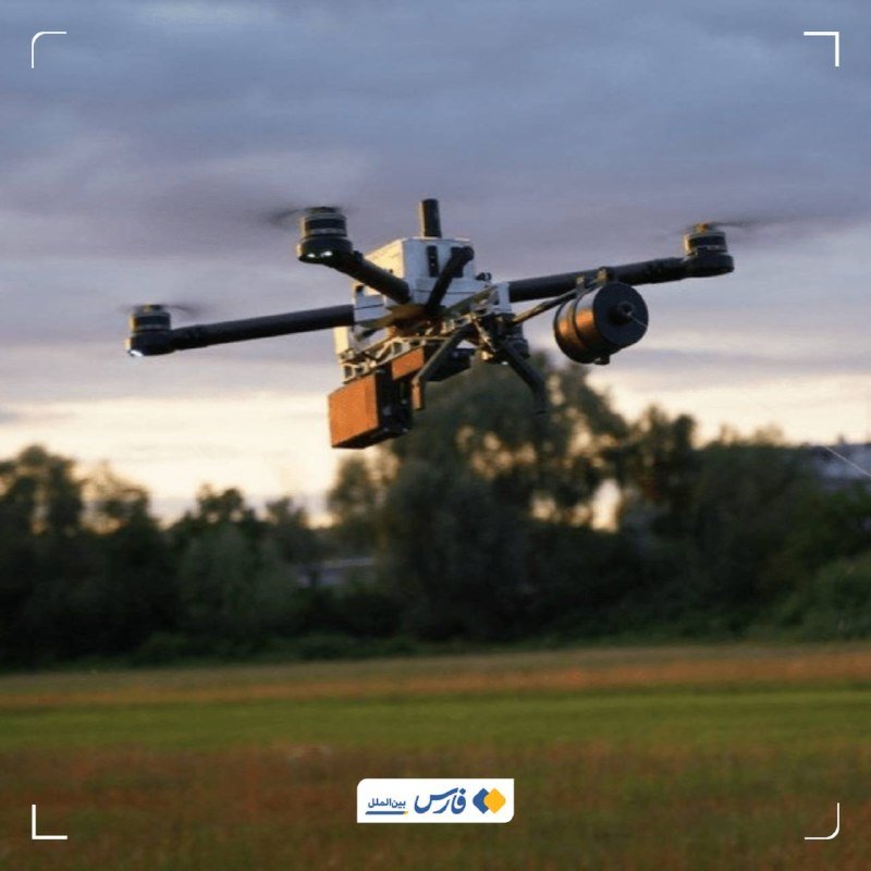 A multi-rotor drone equipped with a cylindrical payload hovers above a grassy field against a cloudy evening sky.