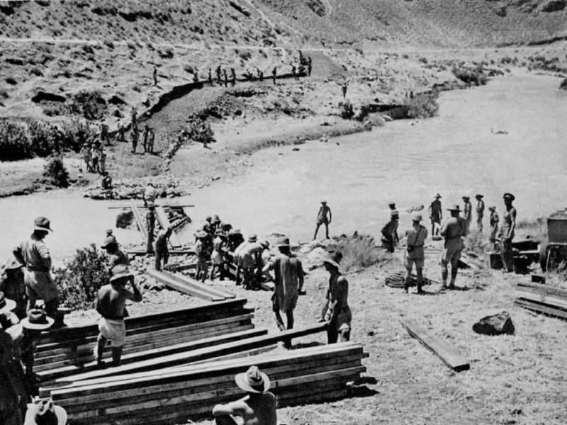 Men in hats work with stacked lumber beside a river, with additional figures visible on the rocky hillside above.