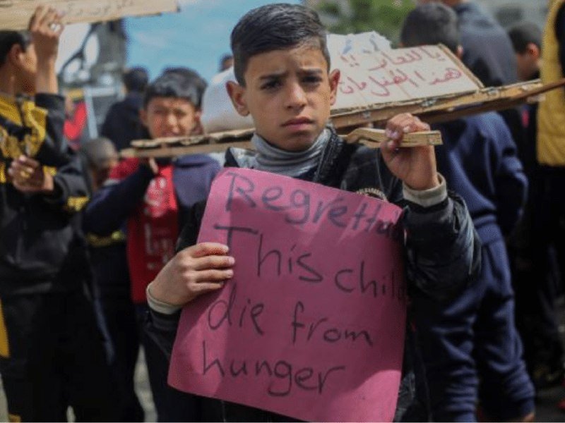 A young boy holds a pink sign reading "Regretted This Child die from hunger" during a crowded outdoor gathering, with other protesters visible behind him holding cardboard signs with Arabic writing.