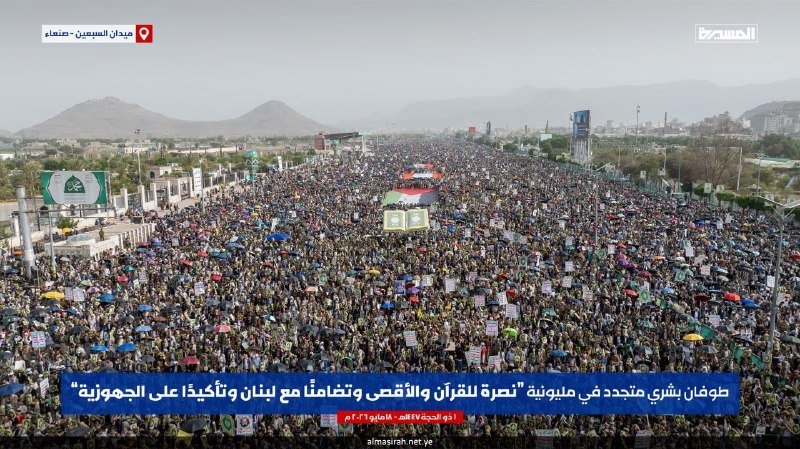 Massive crowd gathered in a city street with mountains in the background, holding signs and banners, with Arabic text overlay indicating Yemen.