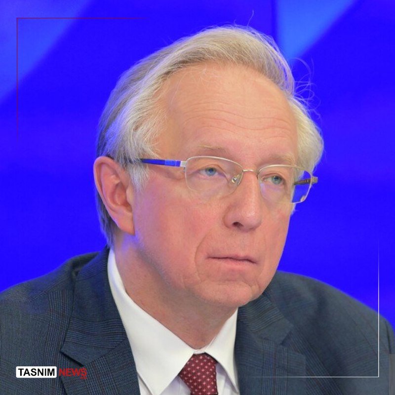 An older man with gray hair, wearing glasses, a gray suit, white shirt, and red patterned tie, sits against a blue background.