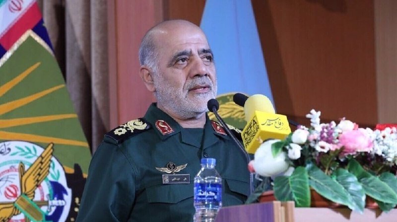 A bearded man in a green military uniform speaks at a podium with microphones, water bottle, and flowers, with an emblem and flag visible behind him.