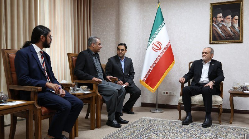 Four men in suits sit in armchairs during a meeting in a formal room featuring an Iranian flag and a framed portrait on the wall.