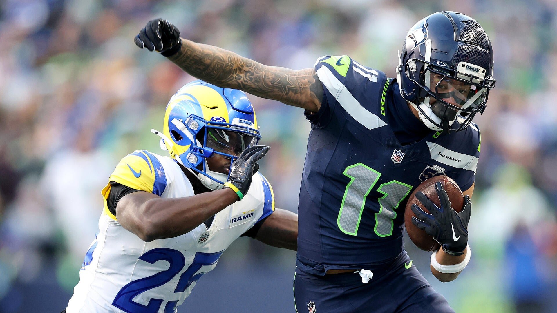 A Seattle Seahawks player in a navy jersey carries a football while stiff-arming a Los Angeles Rams defender in a white jersey during an NFL game.
