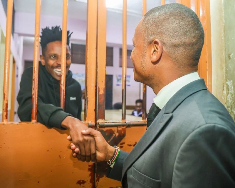 A man in a gray suit shakes hands through orange prison bars with a smiling younger man wearing a black hoodie inside a detention facility.