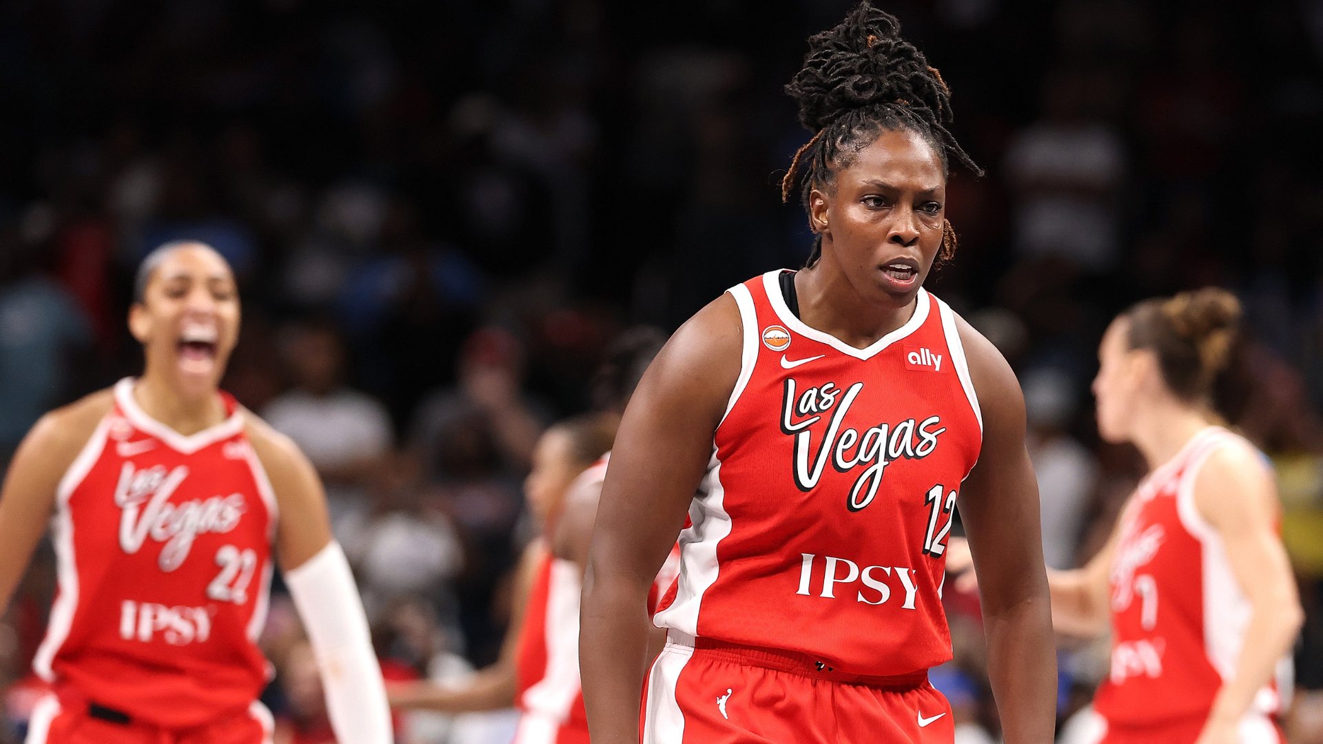 Las Vegas Aces basketball players in red "Las Vegas" jerseys, one shouting in celebration while another wearing number 12 walks past during a game.