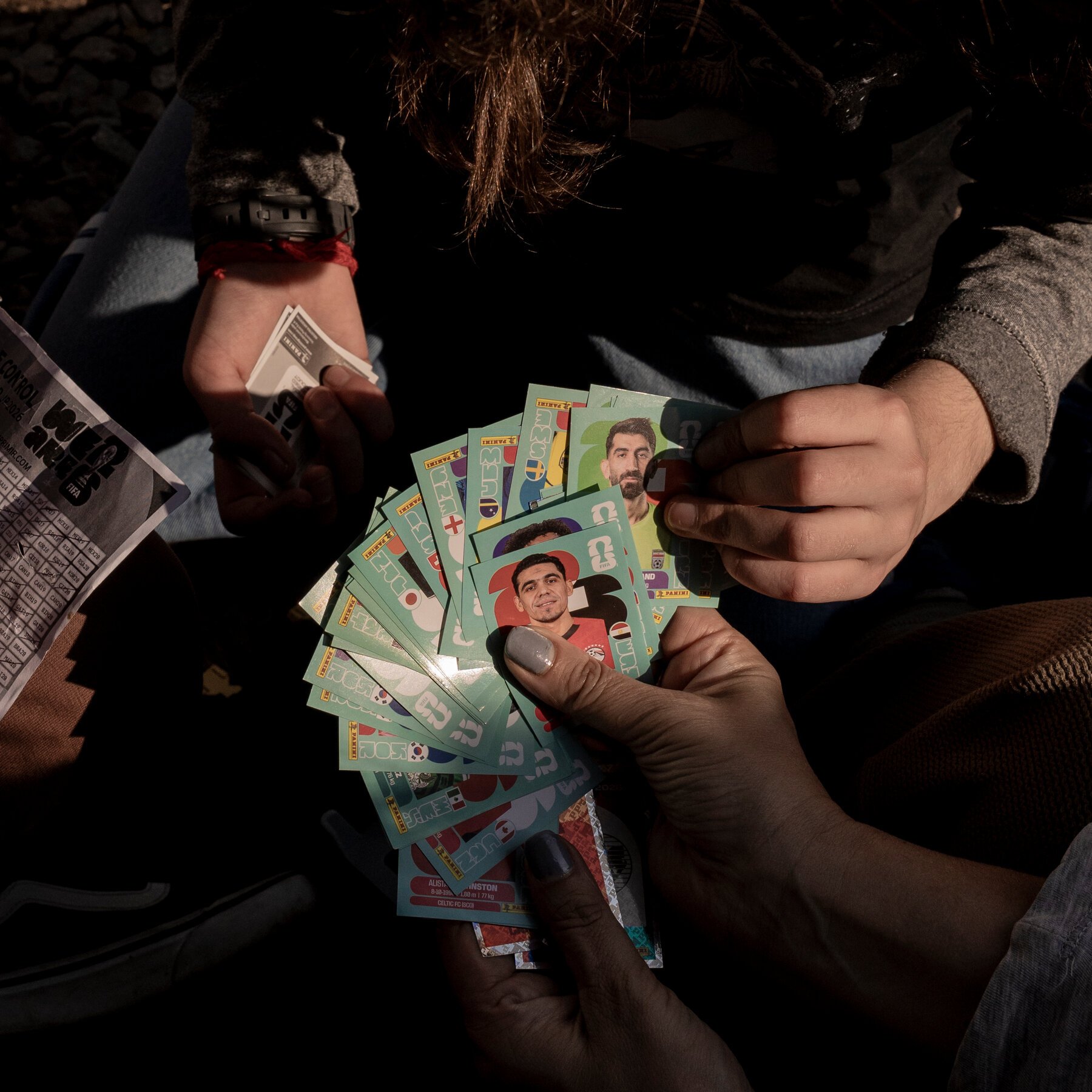 Multiple pairs of hands sort through a fan of Panini soccer trading stickers and cards in a World Cup 2026 album, with a person's head visible above the lap.