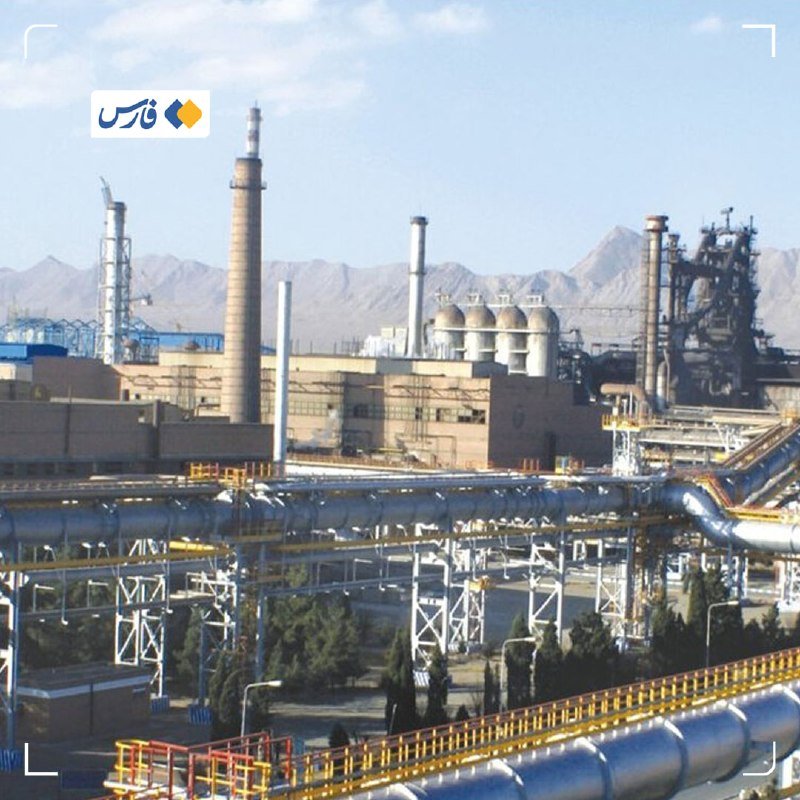 An industrial facility with smokestacks, pipelines, and processing units sits against a backdrop of barren mountains under a hazy sky.